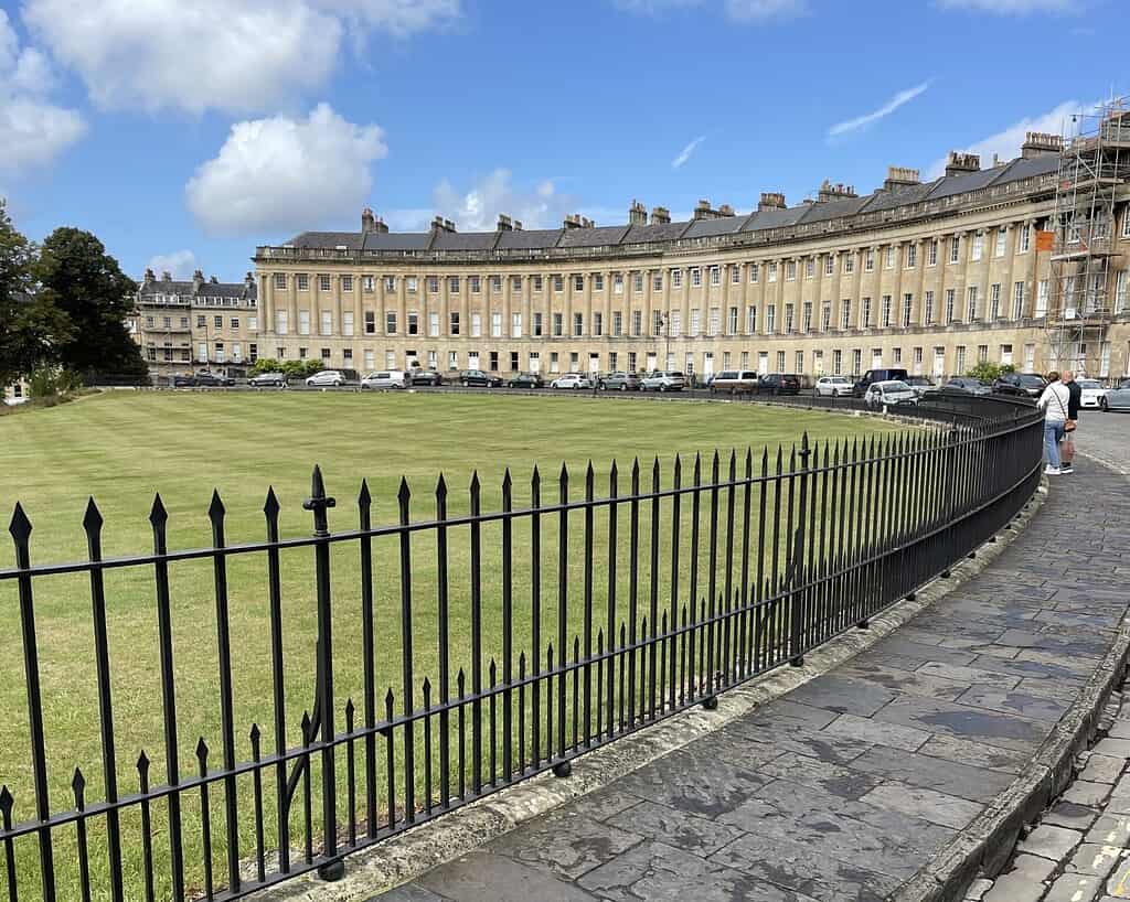 Een gebogen rij historische Georgische huizen staat op de achtergrond, tegenover een groot grasveld omzoomd door een zwart ijzeren hek onder een blauwe hemel. Aan de rechterkant staan twee mensen op de stenen loopbrug - een perfecte stop op elke Jane Austen tour.