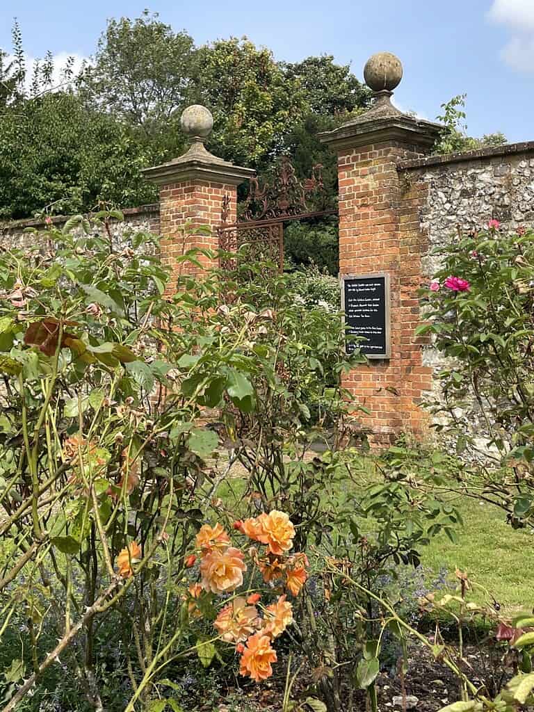 Een tuin met bloeiende oranje en roze rozen op de voorgrond doet denken aan een roman van Jane Austen, terwijl bakstenen pilaren bekroond met stenen bollen en een smeedijzeren hek op de achtergrond staan. Aan een van de pilaren is een bord bevestigd.