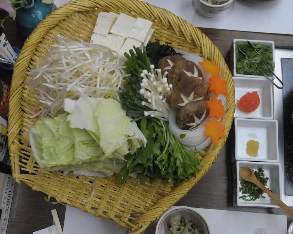 Een bamboemand gevuld met tofu, champignons, taugé, plakjes wortel, kool, bladgroente en enoki-paddenstoelen, omringd door kleine schaaltjes met specerijen op een tafel.