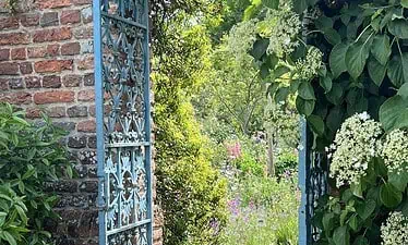 Een stenen pad loopt door een weelderige tuin vol kleurrijke bloemen en groene struiken, die doet denken aan een Kent & Sussex nazomerreis. Gesnoeide heggen en bomen omlijsten het tafereel onder een heldere hemel.