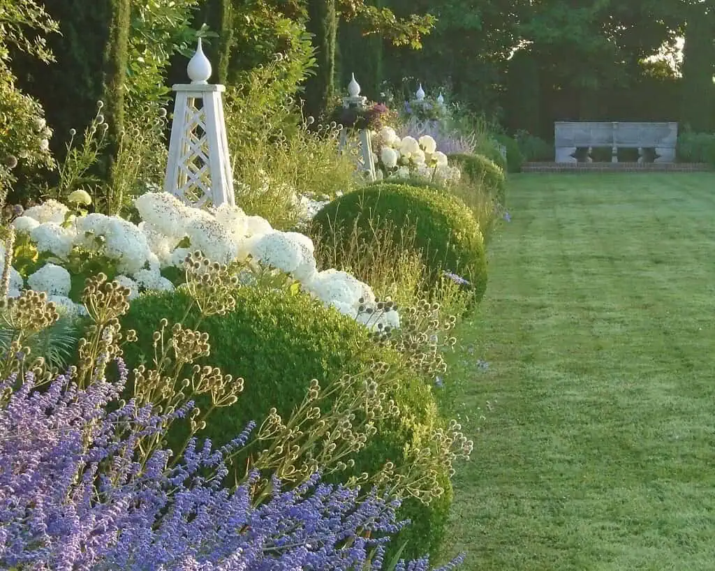 Een keurig verzorgde tuin in Kent heeft weelderig groen gras, bloeiende lavendel, witte hortensia's, geschoren ronde struiken, een wit hekwerk en een stenen bank in de verte - een uitnodigend voorbeeld van klassieke Privétuinen onder gedempt zonlicht.
