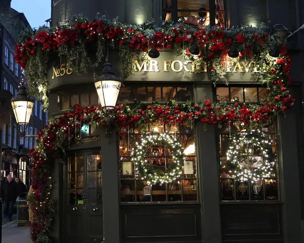 Een traditionele pub in Kent, versierd met rode slingers en gloeiende kransen met de tekst "MR FOGG'S TAVERN" boven de deur. Warme lichtjes en wintersfeer verlichten de feestelijke buitenkant en creëren een gezellige en uitnodigende sfeer in de schemering.