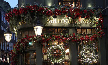 Een traditionele pub in Kent, versierd met rode slingers en gloeiende kransen met de tekst "MR FOGG'S TAVERN" boven de deur. Warme lichtjes en wintersfeer verlichten de feestelijke buitenkant en creëren een gezellige en uitnodigende sfeer in de schemering.