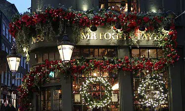 Een traditionele pub in Kent, versierd met rode slingers en gloeiende kransen met de tekst "MR FOGG'S TAVERN" boven de deur. Warme lichtjes en wintersfeer verlichten de feestelijke buitenkant en creëren een gezellige en uitnodigende sfeer in de schemering.