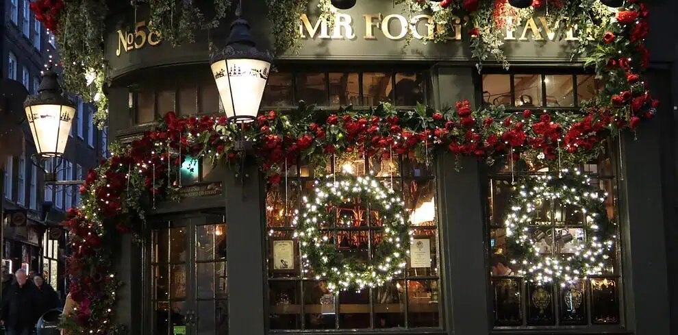 Een traditionele pub in Kent, versierd met rode slingers en gloeiende kransen met de tekst "MR FOGG'S TAVERN" boven de deur. Warme lichtjes en wintersfeer verlichten de feestelijke buitenkant en creëren een gezellige en uitnodigende sfeer in de schemering.