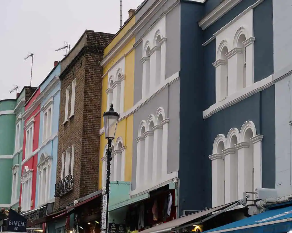 Een rij kleurrijke, ouderwetse gebouwen met witte kozijnen en boogramen roept een charmante Sussex wintersfeer op. De pasteltinten blauw, geel, groen en beige worden aangevuld door een straatlantaarn die ervoor staat.