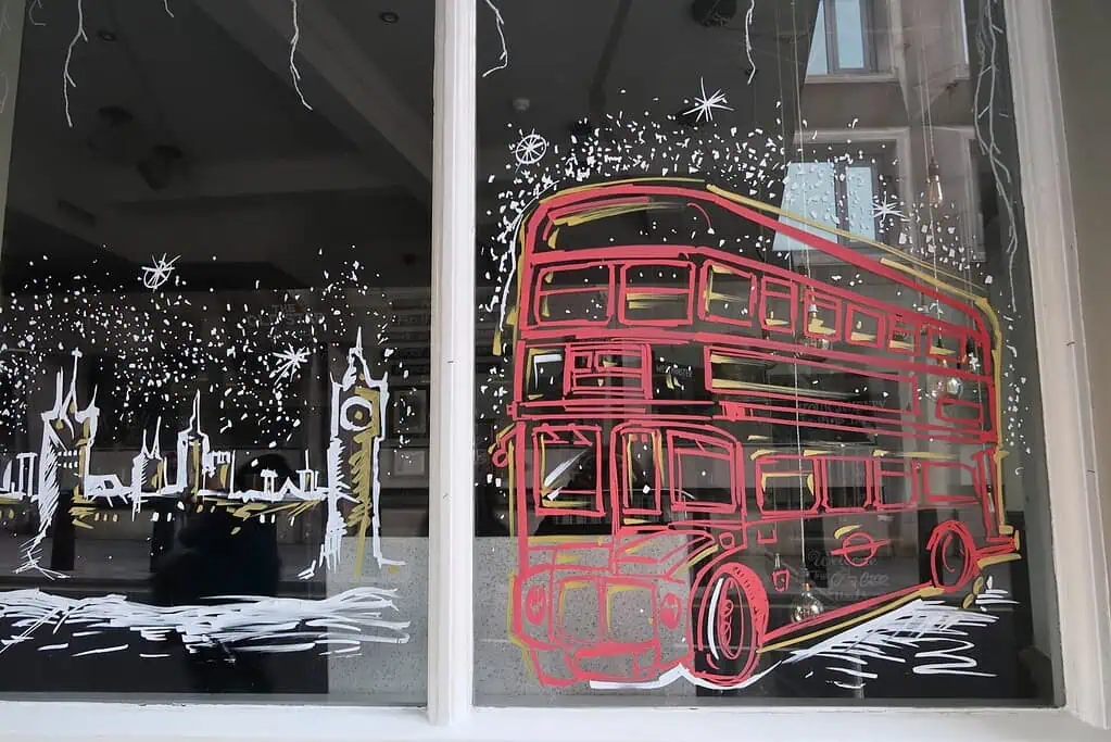 Een raam beschilderd met een rode omtrek van een dubbeldekkerbus en witte schetsen van Londense oriëntatiepunten, waaronder de Big Ben, roept wintersfeer op die doet denken aan Sussex, met sterachtige decoraties die het tafereel omringen.