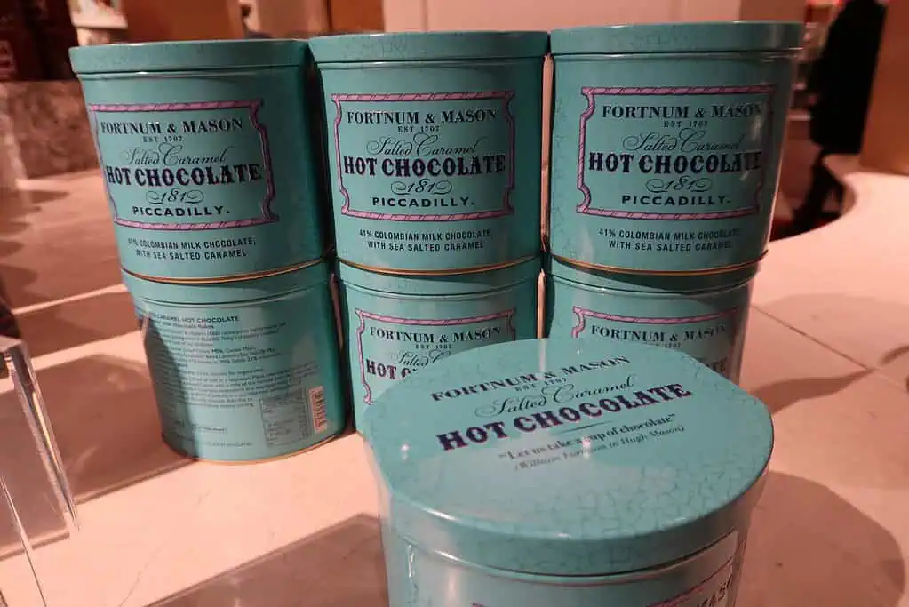 Blikken Fortnum & Mason Salted Caramel Hot Chocolate staan opgestapeld uitgestald, hun groenblauwe verpakking doet denken aan wintersfeer, met blauwe en roze tekst: 45% Colombiaanse melkchocolade met zeezout karamel.