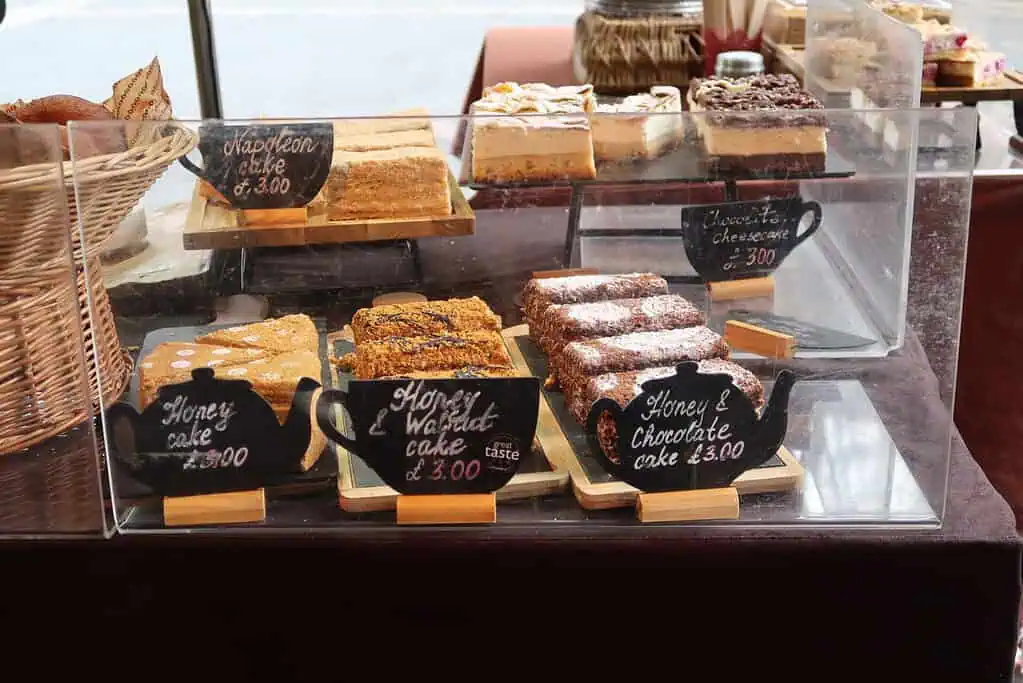 Een bakkerijvitrine met verschillende taarten op dienbladen, waaronder honingcake, honing-walnotentaart en Napoleontaart - elk met een prijsaanduiding op krijtbordbordjes - doet denken aan een gezellige wintersfeer in de patisserieën van Kent of Sussex.