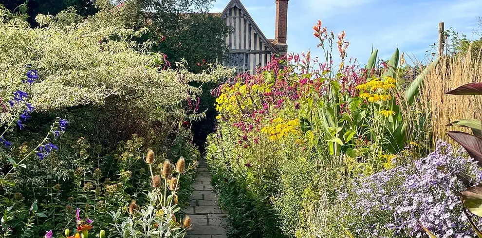 Een stenen pad slingert door een weelderige, kleurrijke tuin vol bloeiende planten en hoge grassen en leidt naar een vakwerkhuis met een bakstenen schoorsteen - perfect voor een zomerreis naar het schilderachtige platteland van Kent of Sussex onder een blauwe hemel.