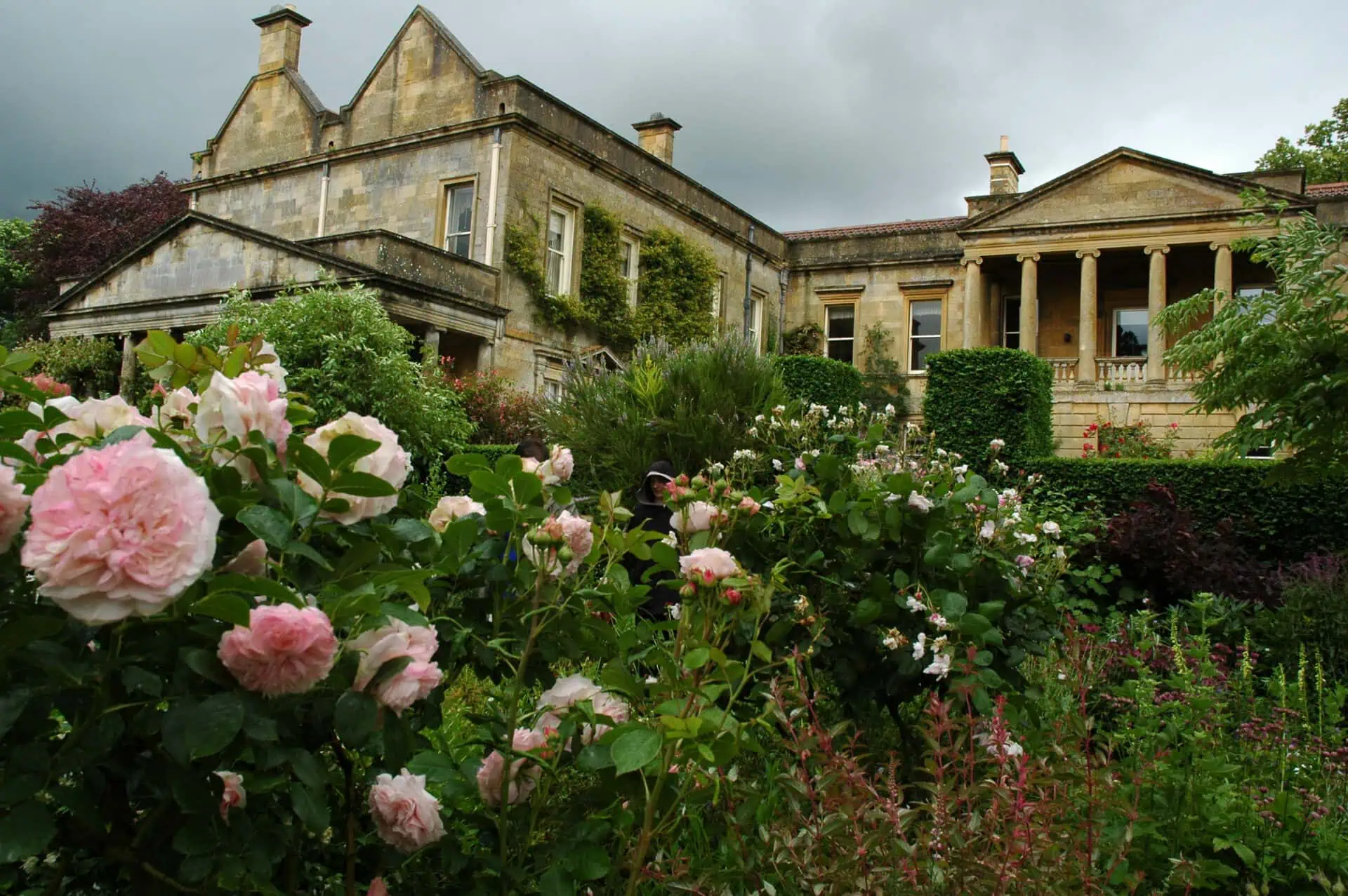 Een groots stenen herenhuis met zuilen en klimop wordt omringd door weelderige tuinen vol bloeiende roze en witte rozen, die doen denken aan de sfeer van de mooiste tuinen van Engeland onder een bewolkte hemel.