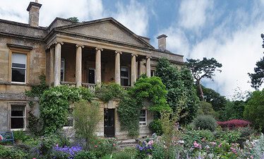 Een historisch stenen herenhuis met hoge zuilen en met klimop bedekte muren staat achter een levendige tuin vol bloeiende roze, paarse en witte bloemen onder een gedeeltelijk bewolkte hemel.