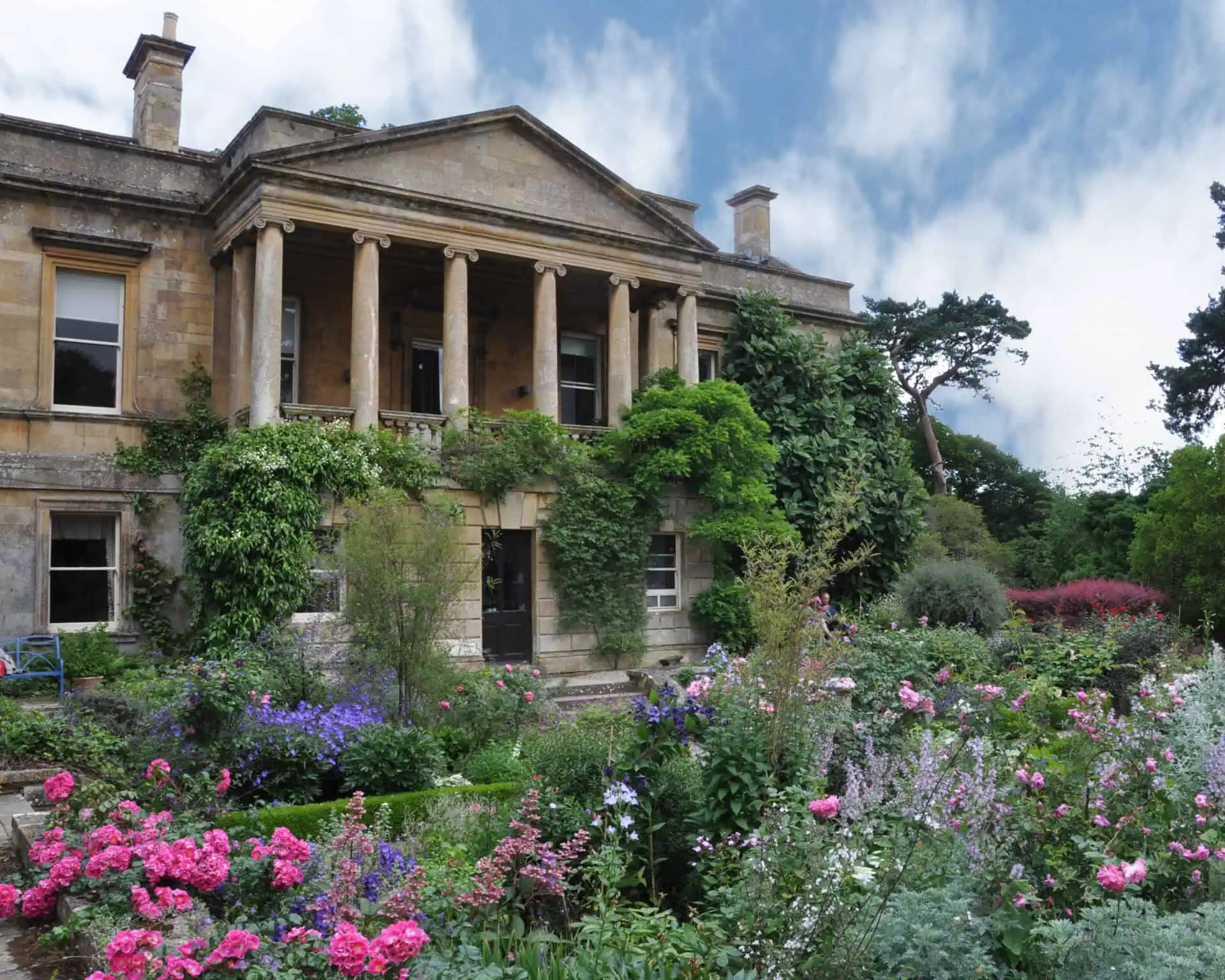 Een historisch stenen herenhuis met hoge zuilen en met klimop bedekte muren staat achter een levendige tuin vol bloeiende roze, paarse en witte bloemen onder een gedeeltelijk bewolkte hemel.