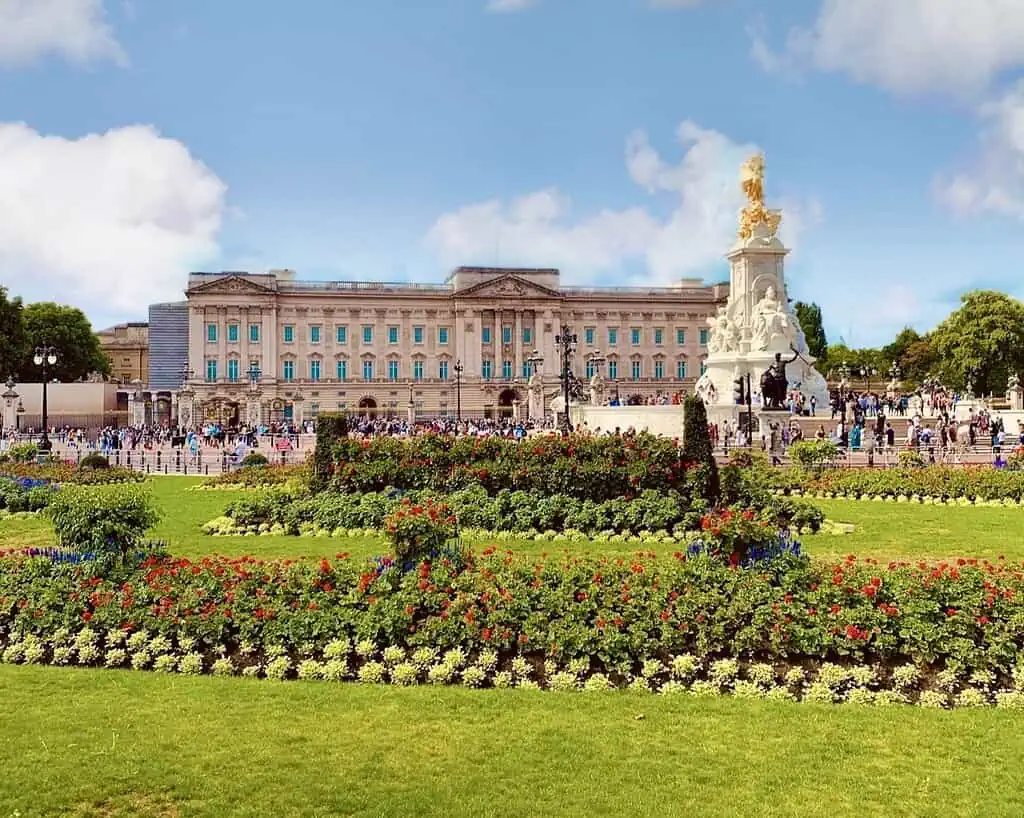 Buckingham Palace met mensenmassa's ervoor, het standbeeld van Victoria Memorial zichtbaar, en kleurrijke bloembedden en groene gazons die doen denken aan koninklijke tuinen onder een gedeeltelijk bewolkte hemel, die een van de grootste paleizen van Londen laat zien.