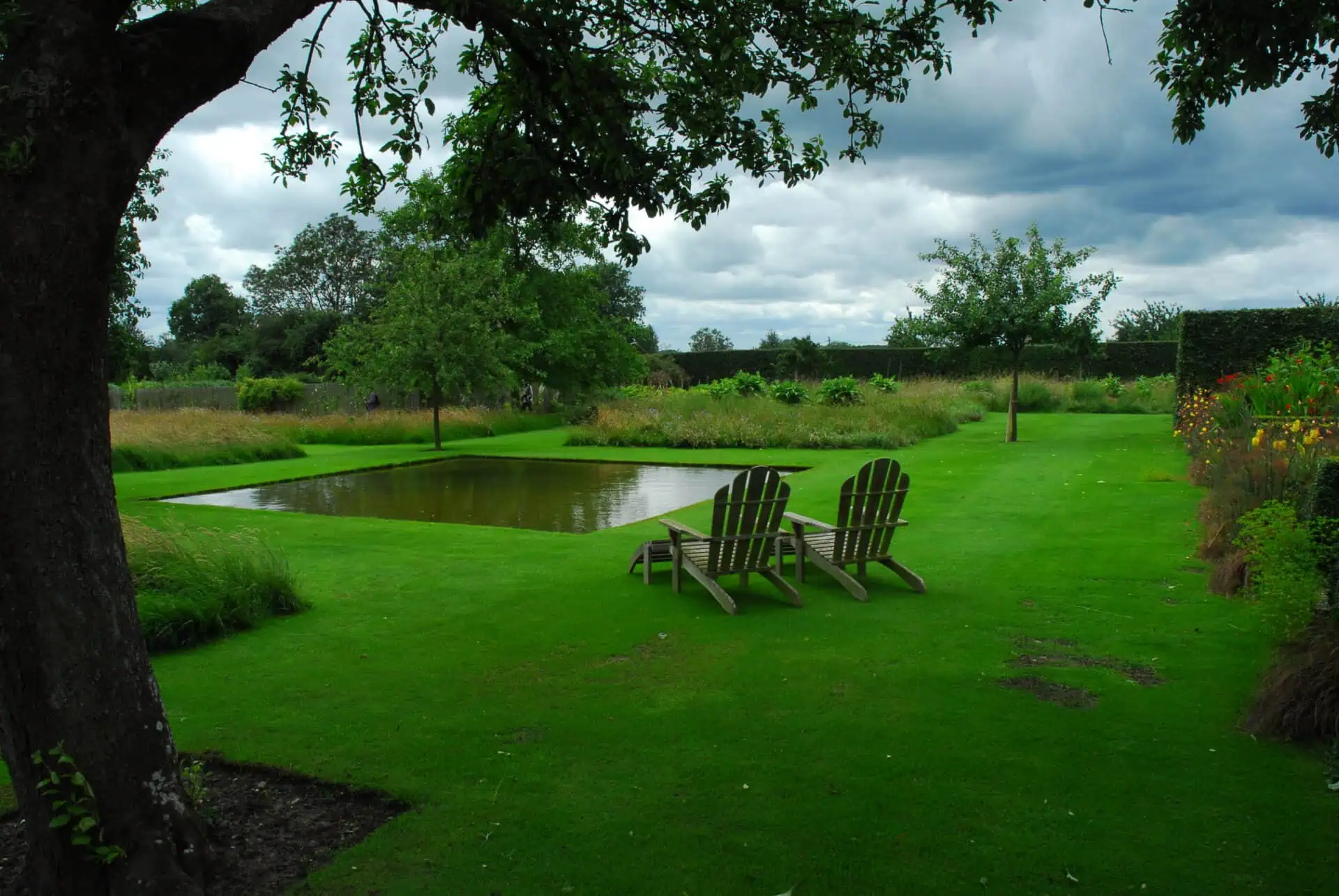 Twee houten stoelen kijken uit op een kleine rechthoekige vijver in een weelderige groene tuin in Le Jardin Plume, omringd door bomen en bloeiende planten onder een bewolkte hemel.