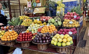 Een kleurrijke marktkraam in de openlucht toont een verscheidenheid aan vers fruit, waaronder sinaasappels, bananen, druiven, granaatappels, drakenfruit en cherimoya's, gerangschikt in manden en stapels met levendige producten rondom.