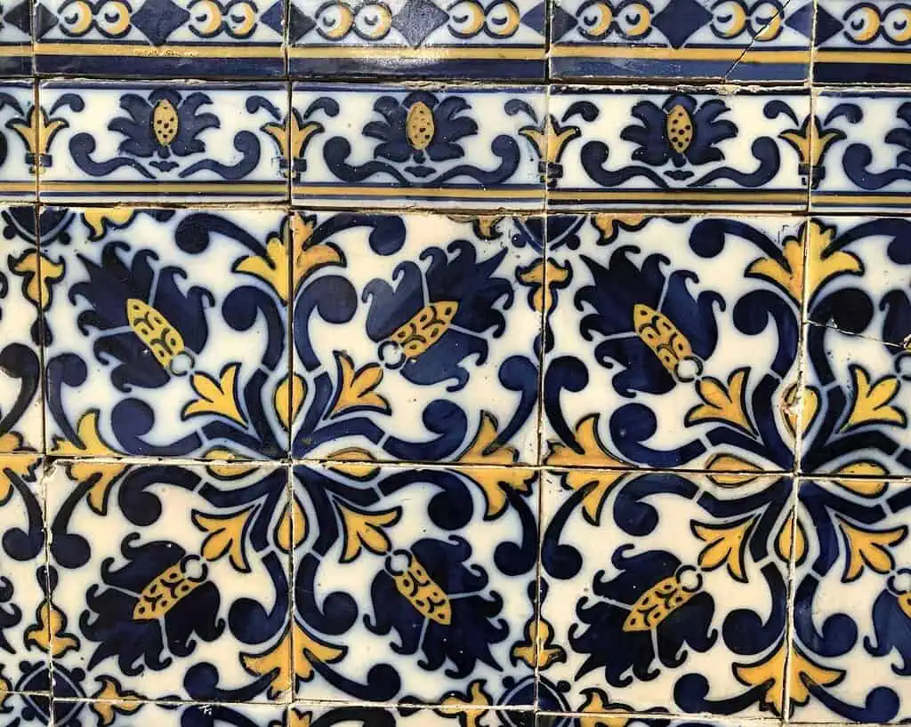 Decoratieve keramische tegels met een symmetrisch blauw, geel en wit bloemen- en geometrisch patroon, met gestileerde bladeren, bloemen en ovale elementen, gerangschikt in een zich herhalend ontwerp. Sommige tegels hebben zichtbare scheuren.