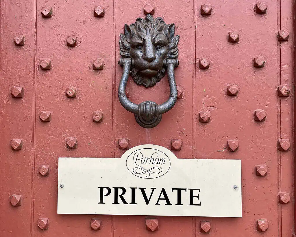 Een rode deur met metalen noppen is voorzien van een decoratieve deurklopper met leeuwenkop boven een wit bord met de tekst "Parham PRIVATE" in zwarte letters, een hint naar het begin van een tuinavontuur geïnspireerd door Marieke Nolsen.