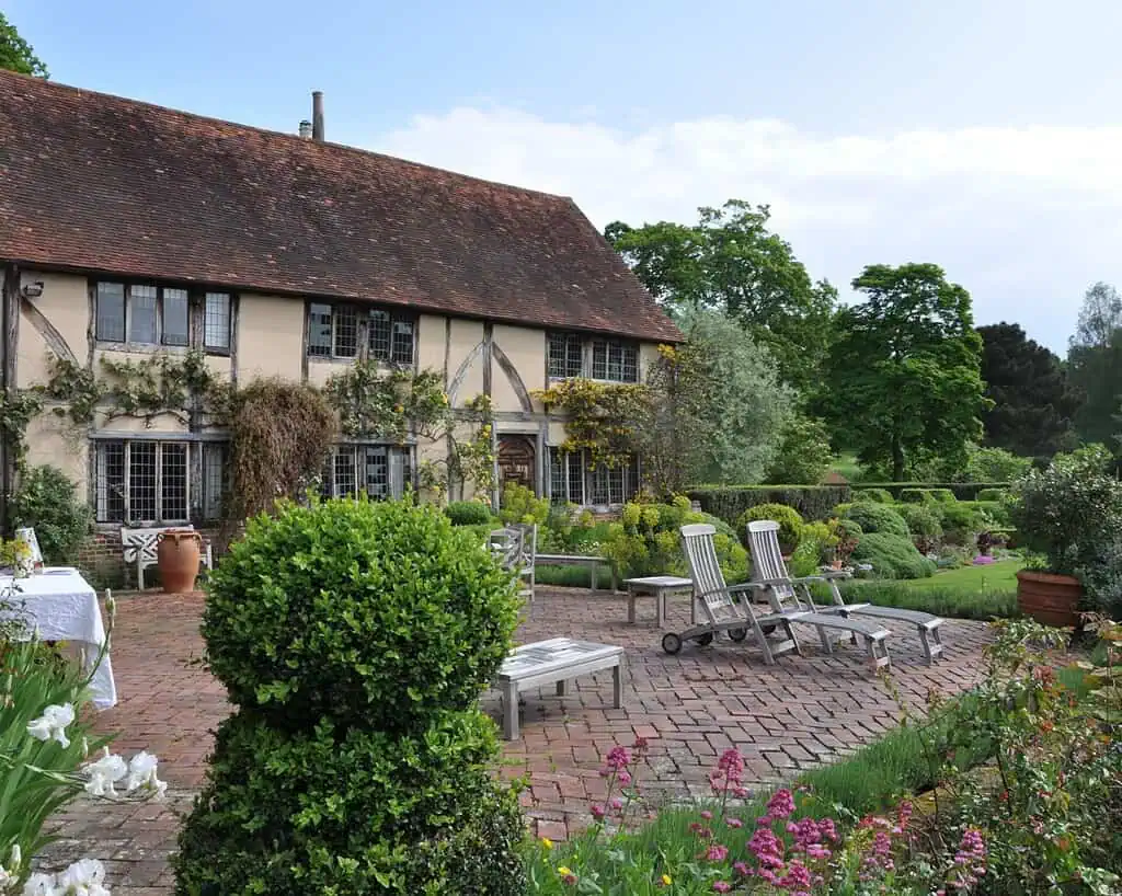 Een charmant oud huis met klimop op de muren ligt achter een bakstenen patio met houten loungestoelen, omringd door weelderig groene struiken en kleurrijke bloembedden - een echt tuinavontuur, zoals Marieke Nolsen zou bewonderen op een zonnige dag.