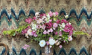 Een groot, kleurrijk boeket roze, paarse en witte bloemen van Marieke Nolsen staat in een zilveren vaas op een houten tafel met een gedessineerde mat, tegen een gedurfd zigzagbehang. Twee stoelen zijn gedeeltelijk zichtbaar aan weerszijden.