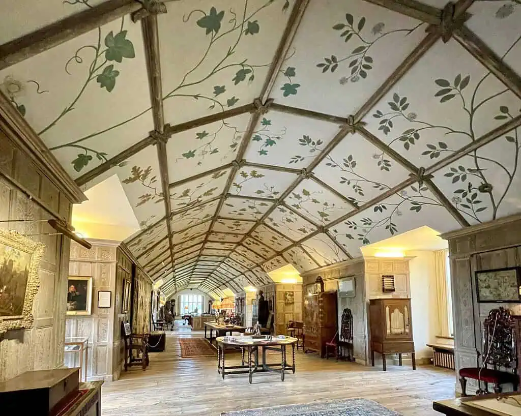 Een lange, elegante hal met een gewelfd plafond met blad- en wijnmotieven, houten vloeren, antieke meubels, schilderijen op wanden met houten panelen en grote ramen die natuurlijk licht binnenlaten - een overdekt tuinavontuur van Marieke Nolsen.