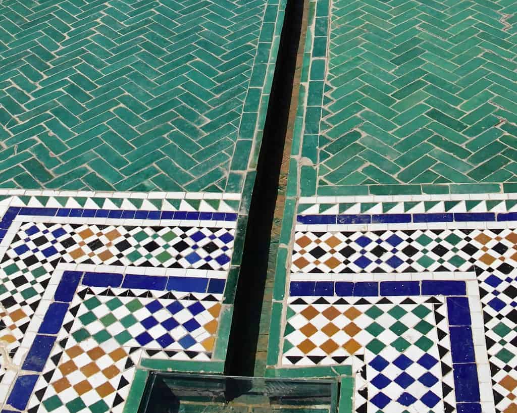 Sierlijke mozaïektegels in groen, blauw, wit, bruin en zwart vormen geometrische patronen met een smal waterkanaal in het midden, van bovenaf gezien.