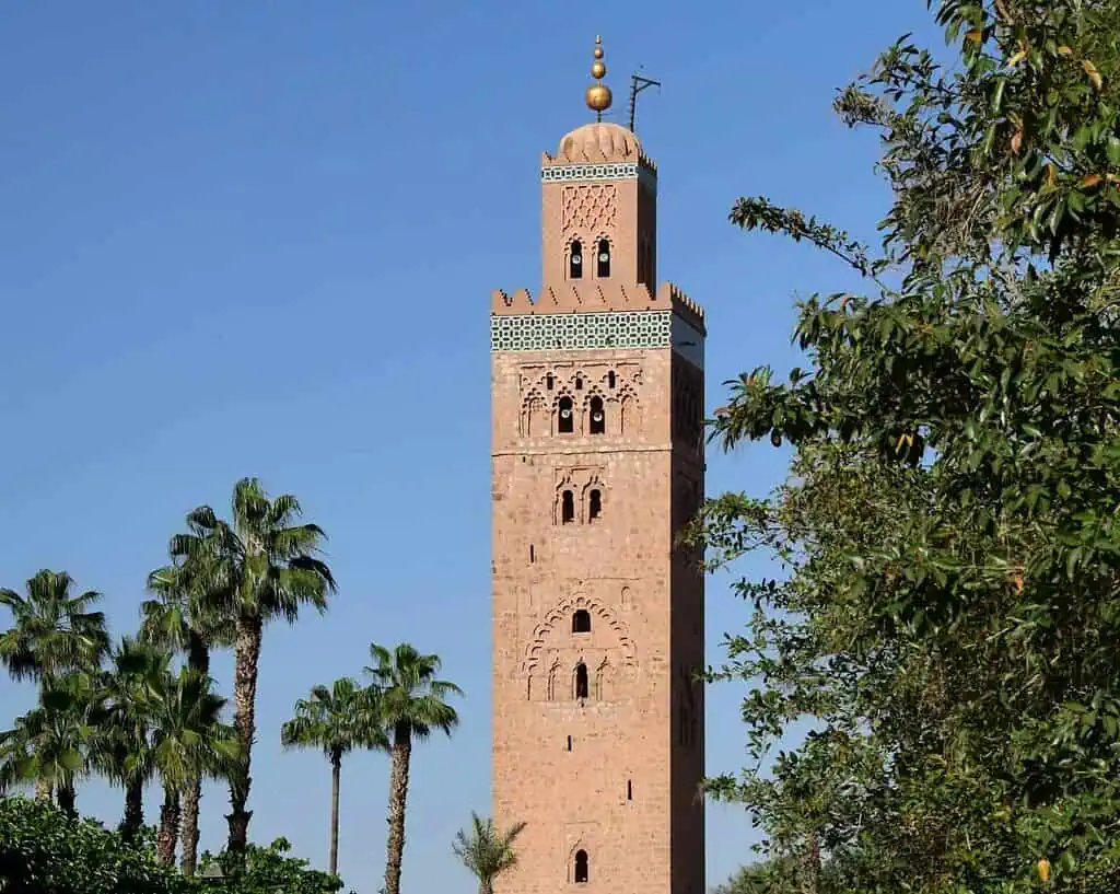 Een hoge, rechthoekige minaret met ingewikkelde patronen, rozeachtige steen en een kleine koepel bovenop staat tegen een helderblauwe hemel, omringd door palmbomen en groen gebladerte.