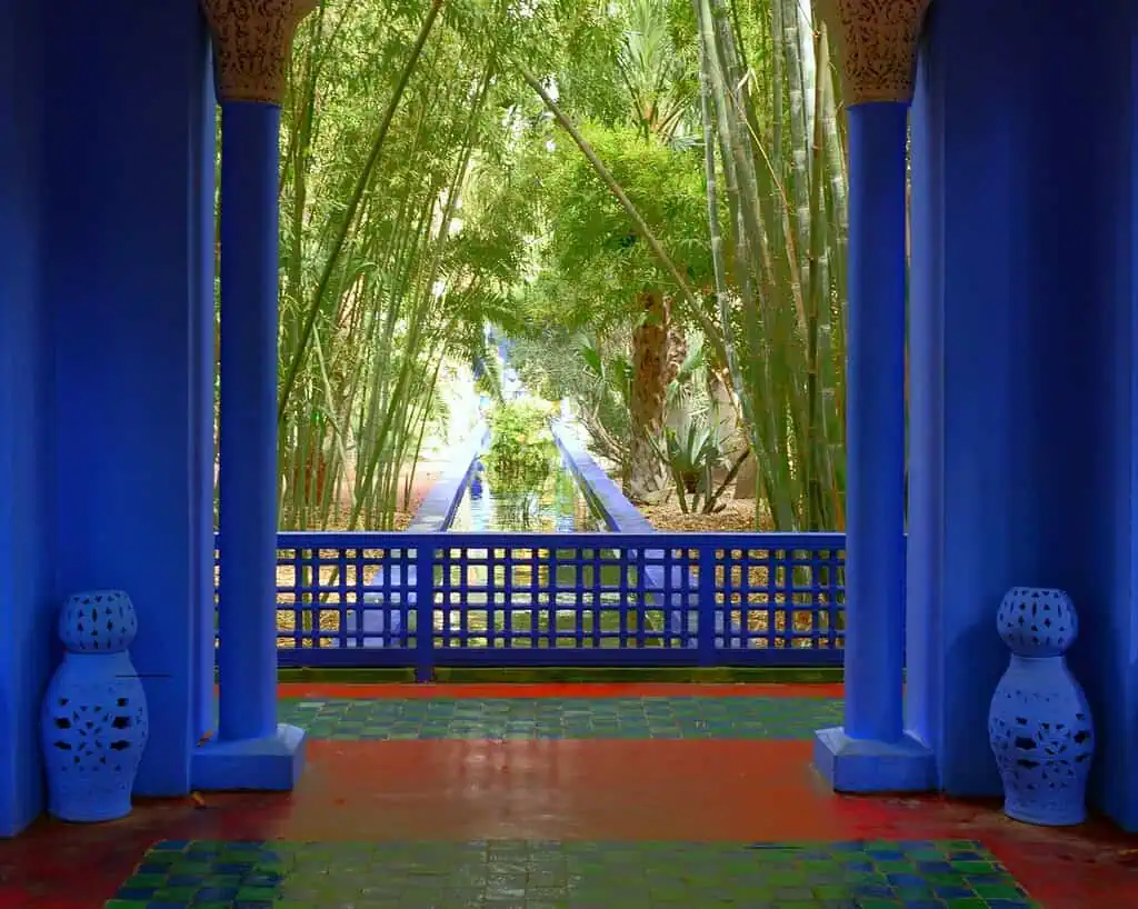 Een uitzicht vanaf een blauw betegelde veranda met zuilen, uitkijkend op een weelderige bamboetuin en een lang, smal spiegelend zwembad in de Majorelle Tuin in Marrakech, Marokko. Twee blauwe decoratieve lantaarns staan naast de zuilen.