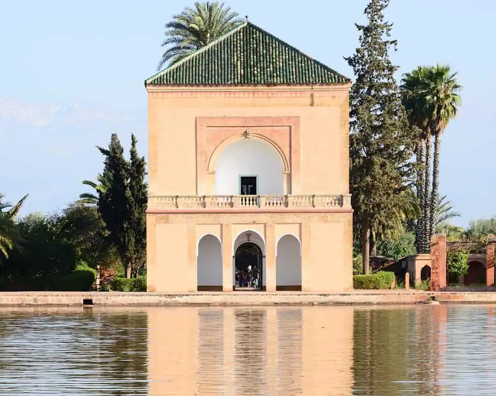 Een historisch paviljoen met een groen pannendak staat naast een rustig spiegelend meer, omringd door palmbomen en cipressen, onder een heldere hemel.