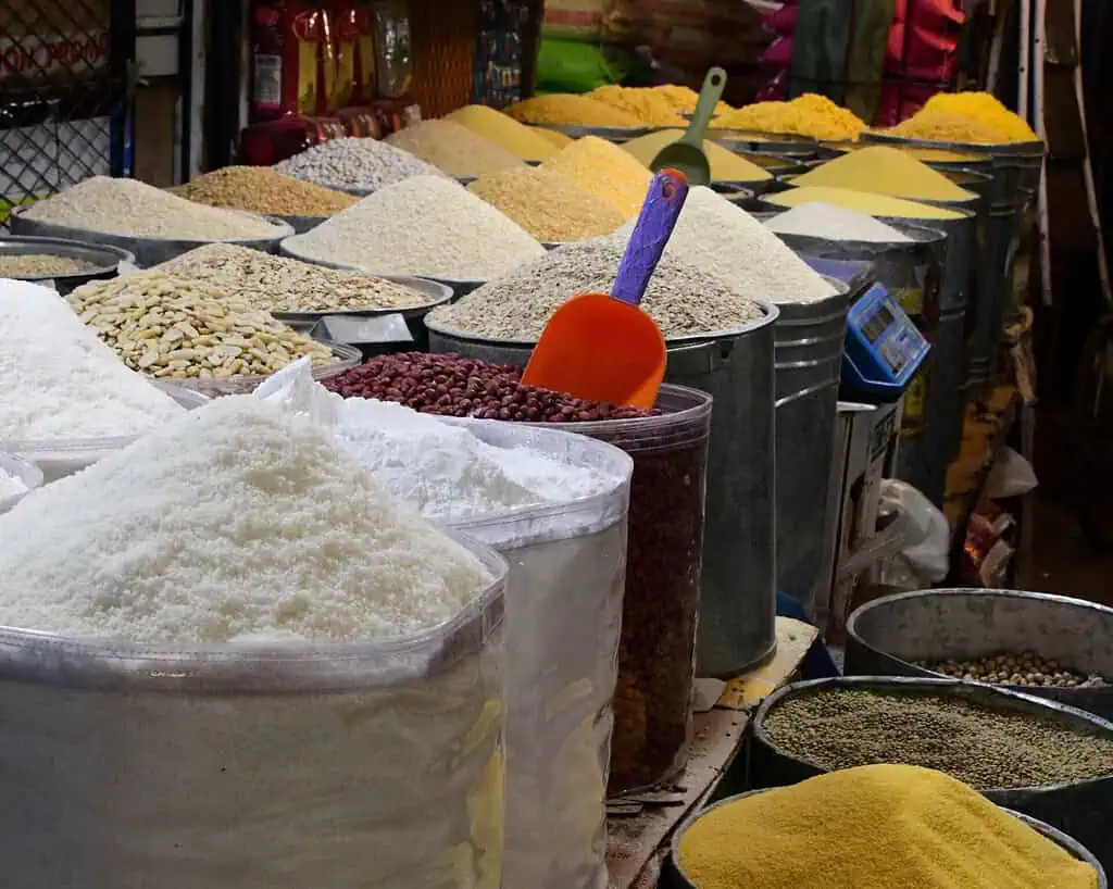 Een assortiment granen, peulvruchten en poeders uitgestald in grote zakken en metalen containers op een markt, met een schep in één container.
