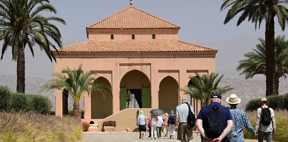 Een groep mensen loopt over een pad met gras en palmbomen in de richting van een bruin gebouw in mediterrane stijl onder een heldere hemel. Sommigen dragen hoeden en tassen en op de achtergrond zijn bergen zichtbaar.