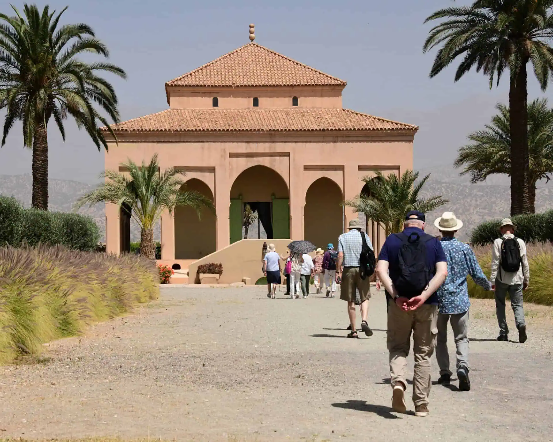 Een groep mensen loopt over een pad met gras en palmbomen in de richting van een bruin gebouw in mediterrane stijl onder een heldere hemel. Sommigen dragen hoeden en tassen en op de achtergrond zijn bergen zichtbaar.