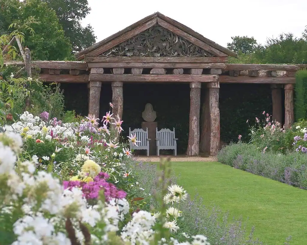 Een tuin met kleurrijke bloemen en een groen gazon leidt naar een rustieke houten pergola met gebeeldhouwde details. Onder de pergola staan twee witte banken en een stenen buste, omringd door weelderig groen.