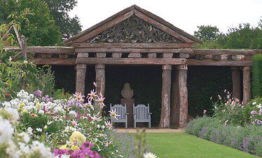 Een tuin met kleurrijke bloemen en een groen gazon leidt naar een rustieke houten pergola met gebeeldhouwde details. Onder de pergola staan twee witte banken en een stenen buste, omringd door weelderig groen.