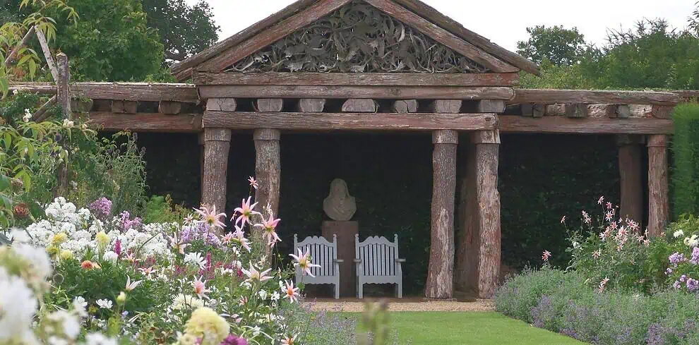 Een tuin met kleurrijke bloemen en een groen gazon leidt naar een rustieke houten pergola met gebeeldhouwde details. Onder de pergola staan twee witte banken en een stenen buste, omringd door weelderig groen.