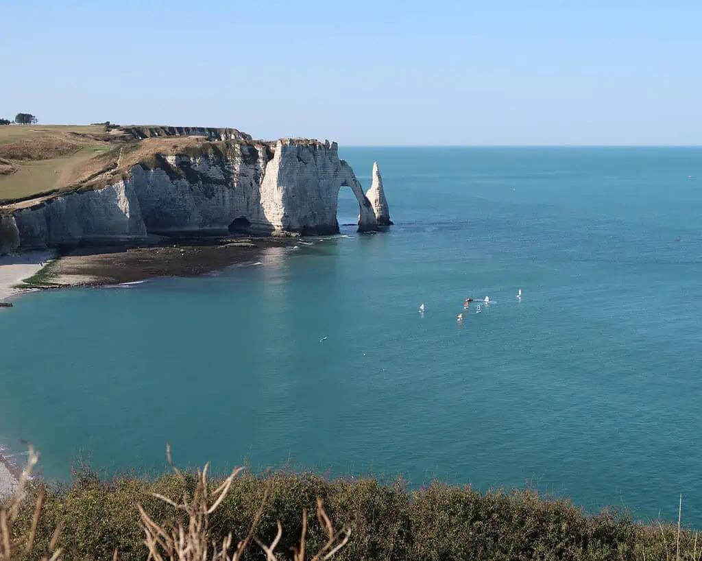 Witte krijtrotsen met een natuurlijke boog rijzen op uit de blauwe zee langs een groene kustlijn. Een paar kleine bootjes of paddleboarders zijn te zien in de buurt van de kliffen onder een heldere, zonnige hemel.