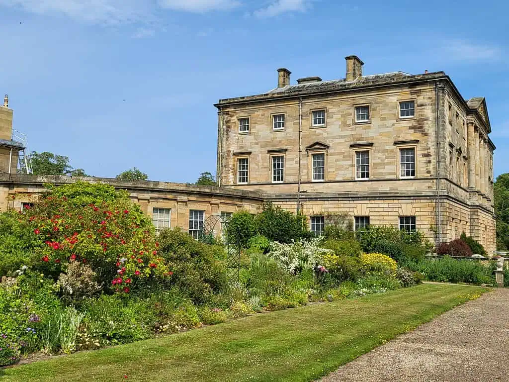 Een groot historisch stenen gebouw met rechthoekige ramen staat naast een weelderige tuin vol kleurrijke bloemen en groene struiken onder een heldere hemel van Northumberland.