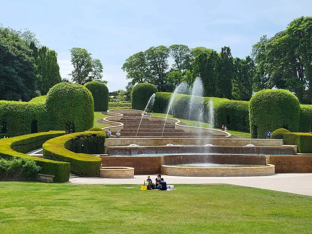 Een trapsgewijs oplopende waterfontein in Northumberland stort zich neer op aangelegde terrassen met keurig gesnoeide heggen en groene bomen. Mensen zitten op het gras en lopen rond in de tuin op een zonnige dag.