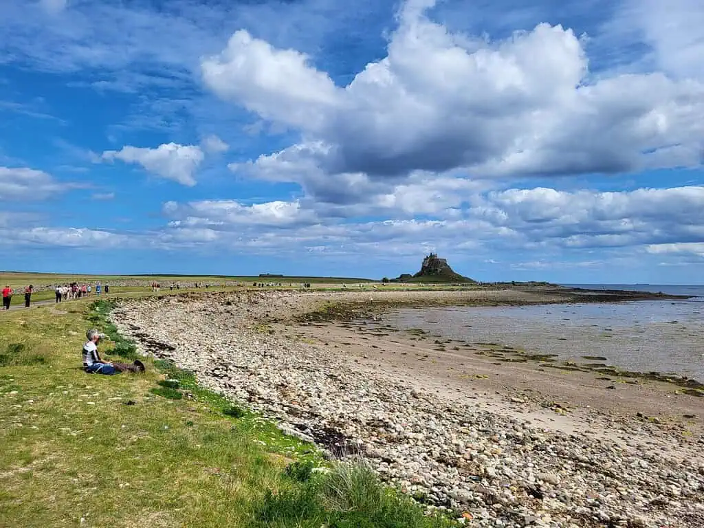 Mensen lopen langs een rotsachtige kustlijn van Northumberland onder een blauwe hemel met verspreide wolken. In de verte staat een historisch kasteel bovenop een kleine heuvel op een eiland. Bij eb zie je nat zand en zeewier langs de kust.
