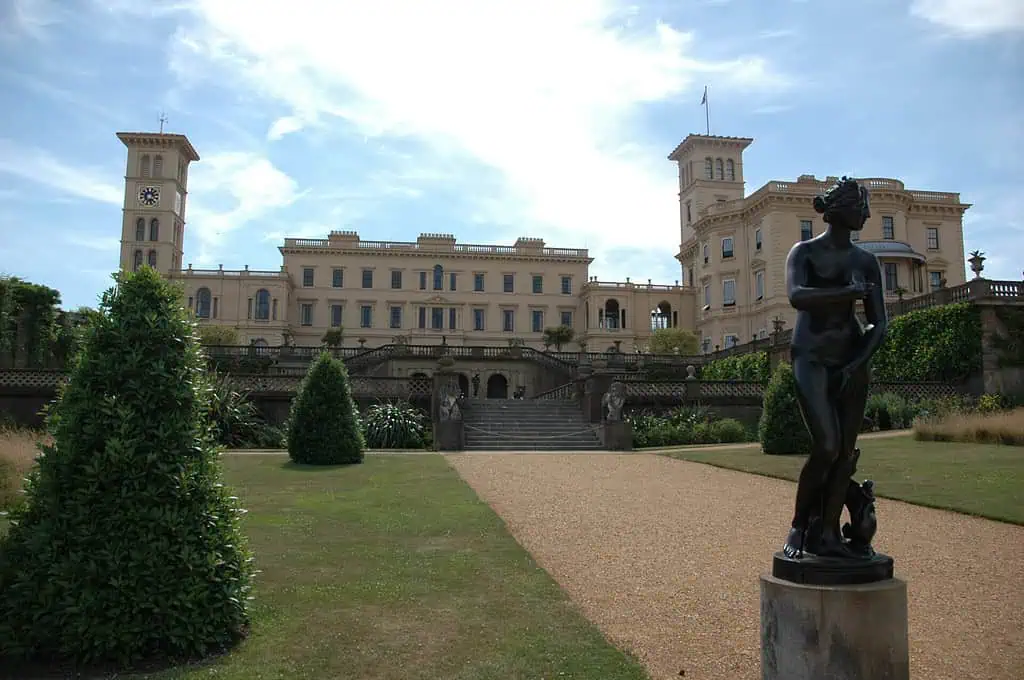 Een groot, crèmekleurig herenhuis dat doet denken aan Osborne House met twee torens staat op de achtergrond, terwijl verzorgde tuinen, struiken en een bronzen vrouwenbeeld op de voorgrond staan onder een gedeeltelijk bewolkte hemel.
