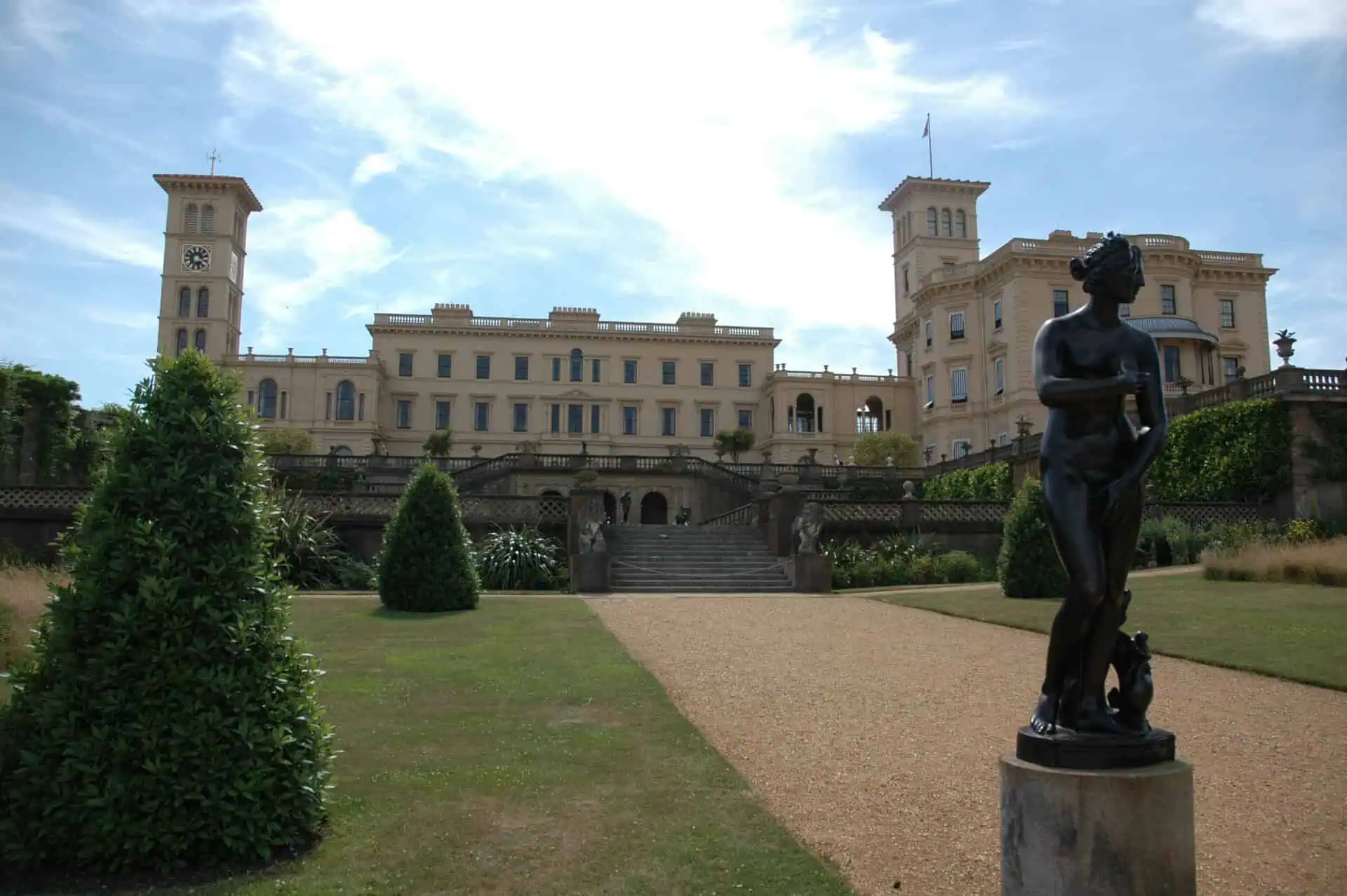 Een groot, crèmekleurig herenhuis dat doet denken aan Osborne House met twee torens staat op de achtergrond, terwijl verzorgde tuinen, struiken en een bronzen vrouwenbeeld op de voorgrond staan onder een gedeeltelijk bewolkte hemel.