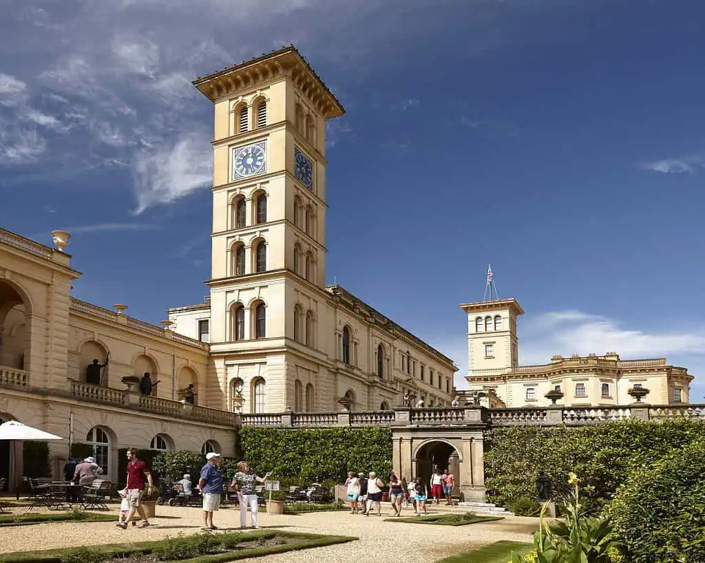Mensen lopen in een tuin naast een groot, sierlijk, crèmekleurig gebouw met een hoge klokkentoren op het Isle of Wight. Het gebouw heeft bogen, zuilen en gedetailleerde architecturale elementen onder een blauwe hemel met pluizige wolken.