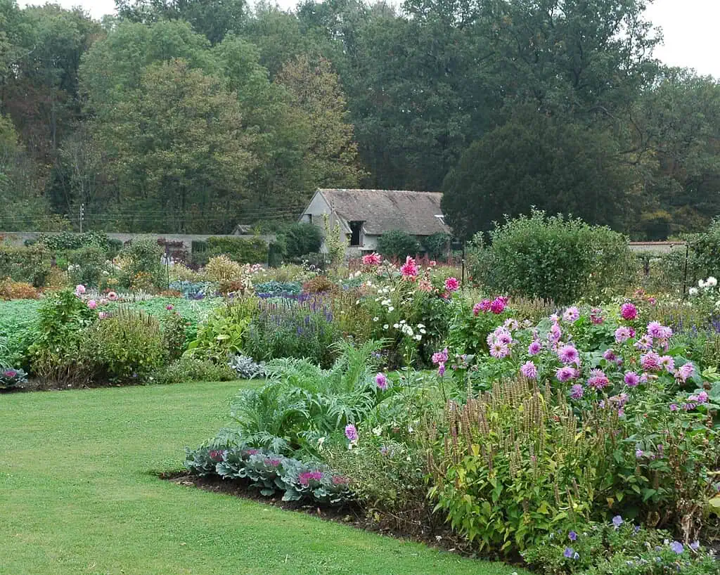 Een weelderige tuin met levendige roze, paarse en witte bloemen, groene struiken en bladplanten, met een klein rustiek schuurtje en dichte bomen op de achtergrond onder een bewolkte hemel.