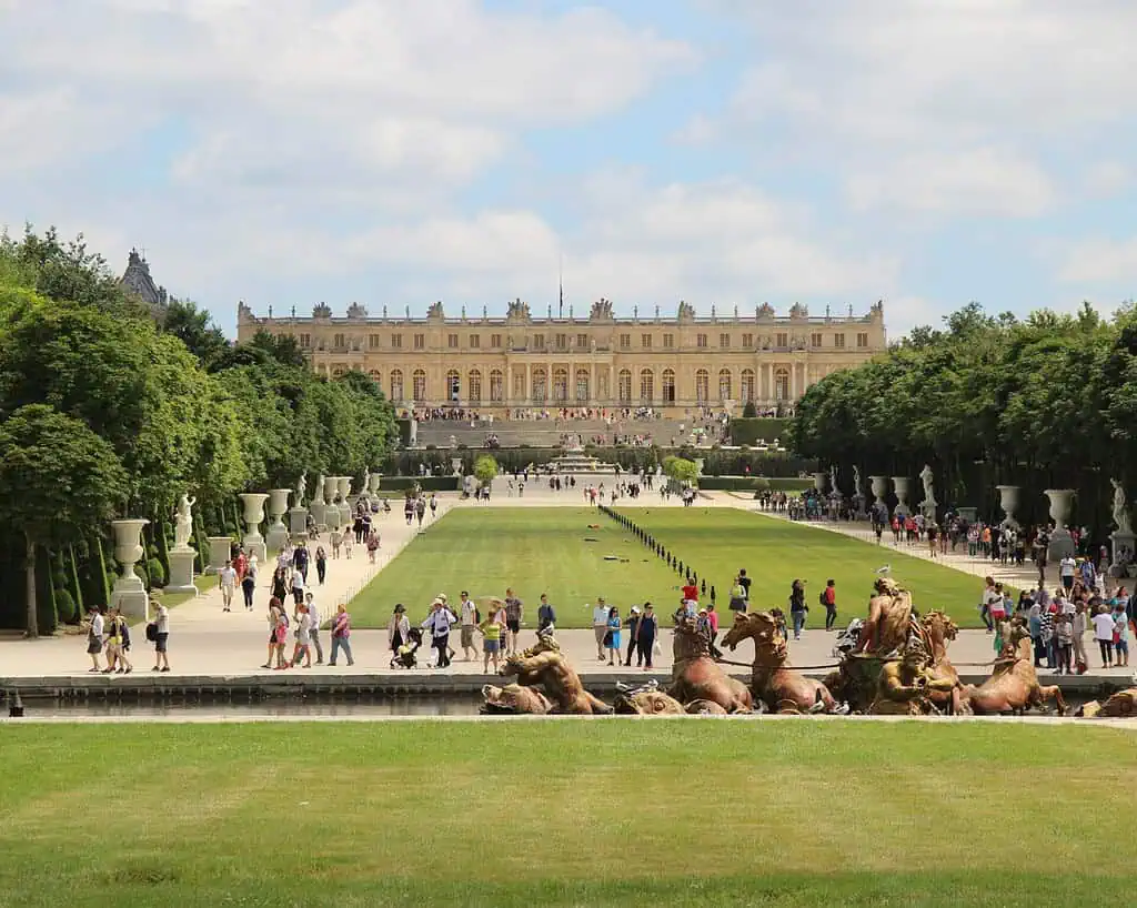 Toeristen wandelen in de tuinen van het paleis van Versailles, met getrimde gazons, standbeelden en een grote fontein op de voorgrond. Het grote paleisgebouw staat op de achtergrond onder een gedeeltelijk bewolkte hemel.