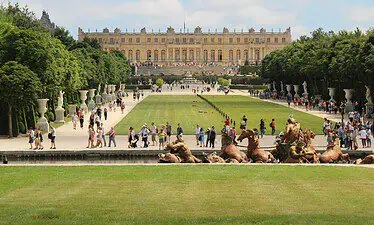 Toeristen wandelen in de tuinen van het paleis van Versailles, met getrimde gazons, standbeelden en een grote fontein op de voorgrond. Het grote paleisgebouw staat op de achtergrond onder een gedeeltelijk bewolkte hemel.