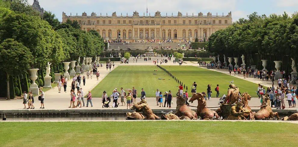 Toeristen wandelen in de tuinen van het paleis van Versailles, met getrimde gazons, standbeelden en een grote fontein op de voorgrond. Het grote paleisgebouw staat op de achtergrond onder een gedeeltelijk bewolkte hemel.