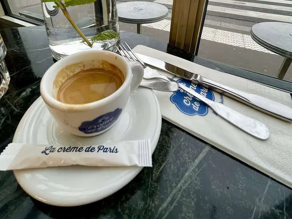 Een klein kopje espresso op een schoteltje staat op een marmeren tafel naast een pakje suiker met de naam La crème de Paris, keukengerei, een servet en een glas water met een munttakje, bij een raam met uitzicht op de straat.