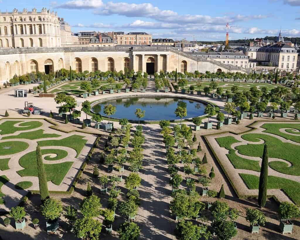 Formele tuinen met symmetrische patronen, rijen bomen in potten en een grote ronde vijver voor een sierlijk paleis met bogen en zuilen onder een blauwe hemel met verspreide wolken.
