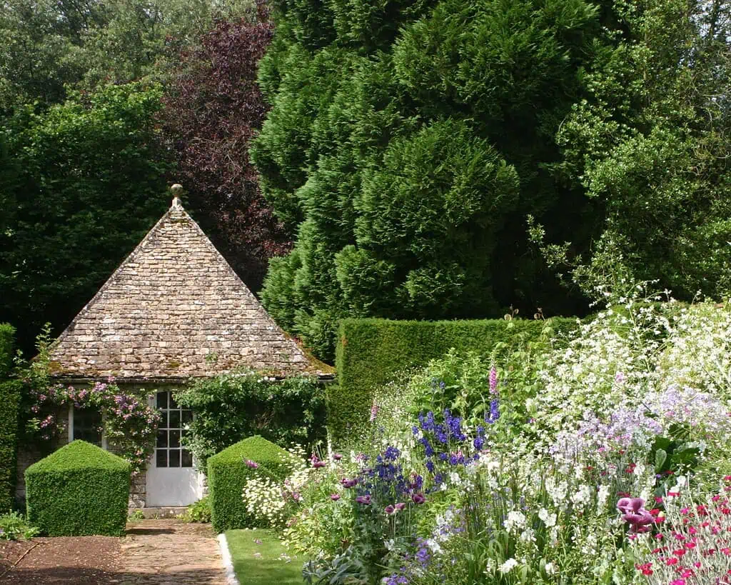 Een klein stenen huisje met een steil schindeldak staat tussen weelderig groen en kleurrijke bloeiende bloemen, omringd door hoge bomen en verzorgde struiken in een levendige tuin.