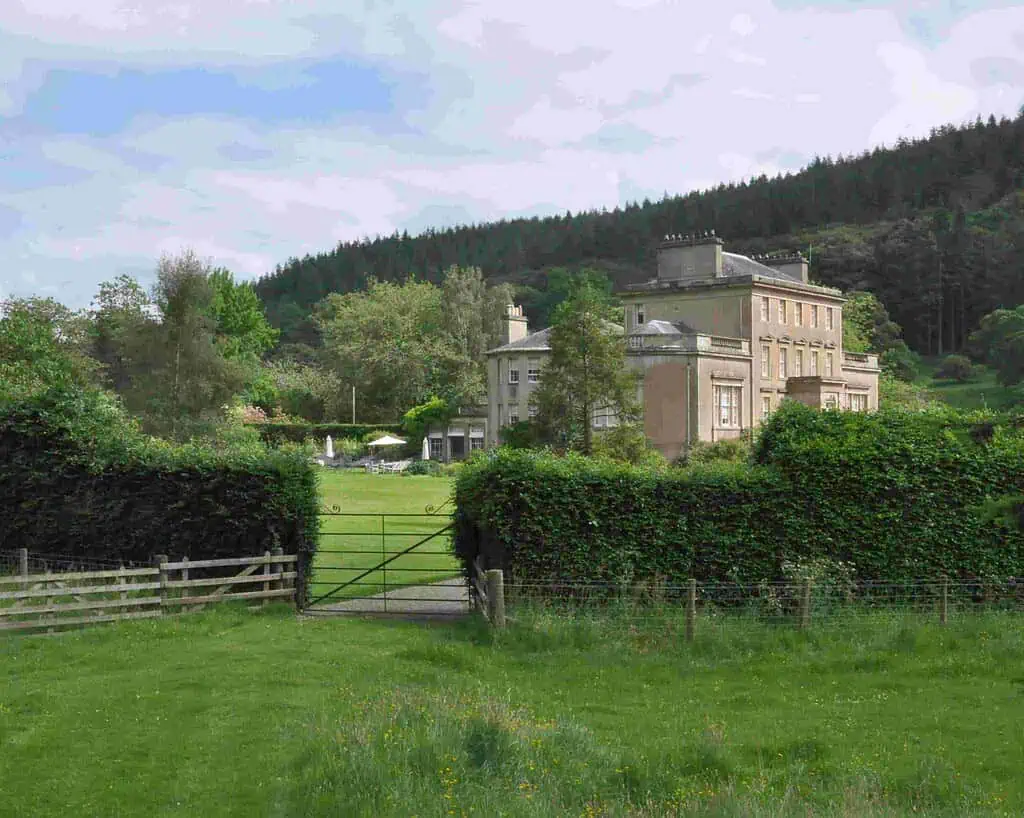 Een groot landhuis met hoge ramen staat achter een heg en een metalen hek, omringd door privétuinen, groene gazons, bomen en beboste heuvels op de achtergrond onder een gedeeltelijk bewolkte Oost-Schotlandse lucht.
